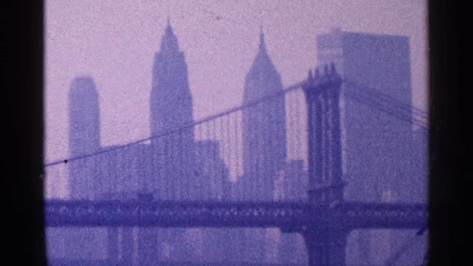 1964 New York City frame showing a suspension bridge over the river with Manhattan skyscrapers beyond it.