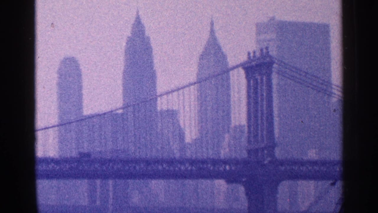 1964 8mm view of New York City skyline with the Manhattan Bridge, distant skyscrapers, and soft haze over the waterfront.