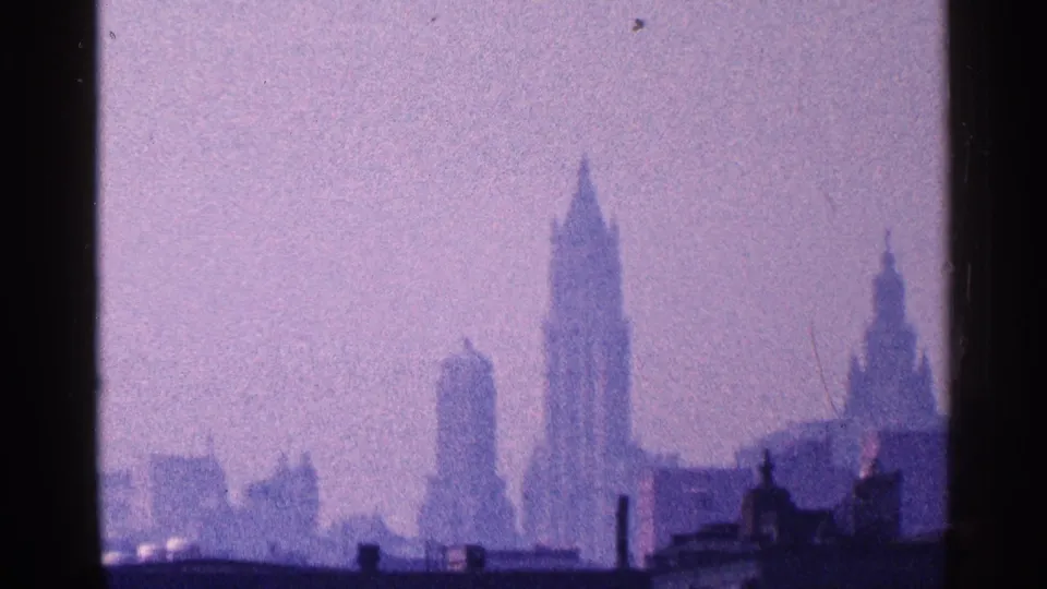 1964 8mm frame of Manhattan skyscrapers and low waterfront structures, softened by haze and purple-tinted film color.