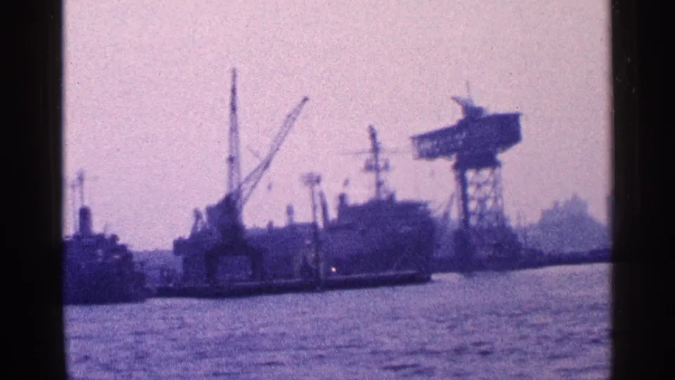 New York City waterfront in 1964 with industrial cranes, docked ships, a large cargo vessel, and rippled water in the foreground.