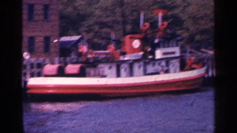 Red-and-white tugboat at a New York City waterfront dock in 1964, with brick building, trees, and water visible.