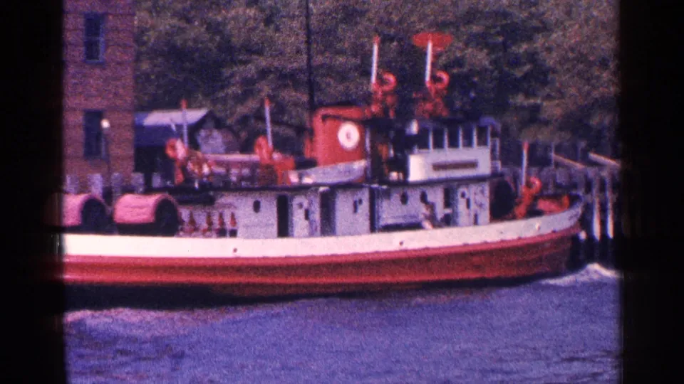 Close side view of the docked red-and-white tugboat at the 1964 New York City waterfront with water and trees behind.