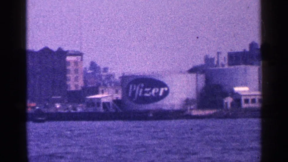 1964 New York City waterfront frame showing a large Pfizer sign on an industrial building across the river.
