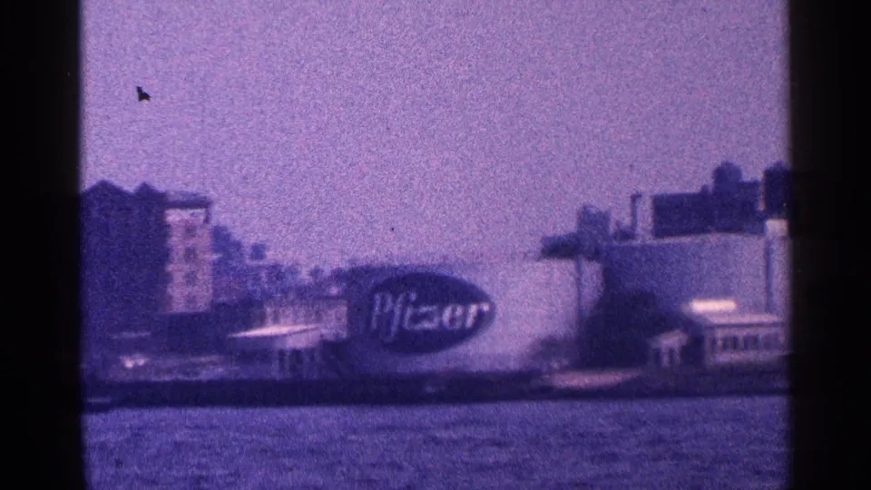 New York City harbor-tour view with the Pfizer sign and waterfront buildings visible through grainy 8mm film.