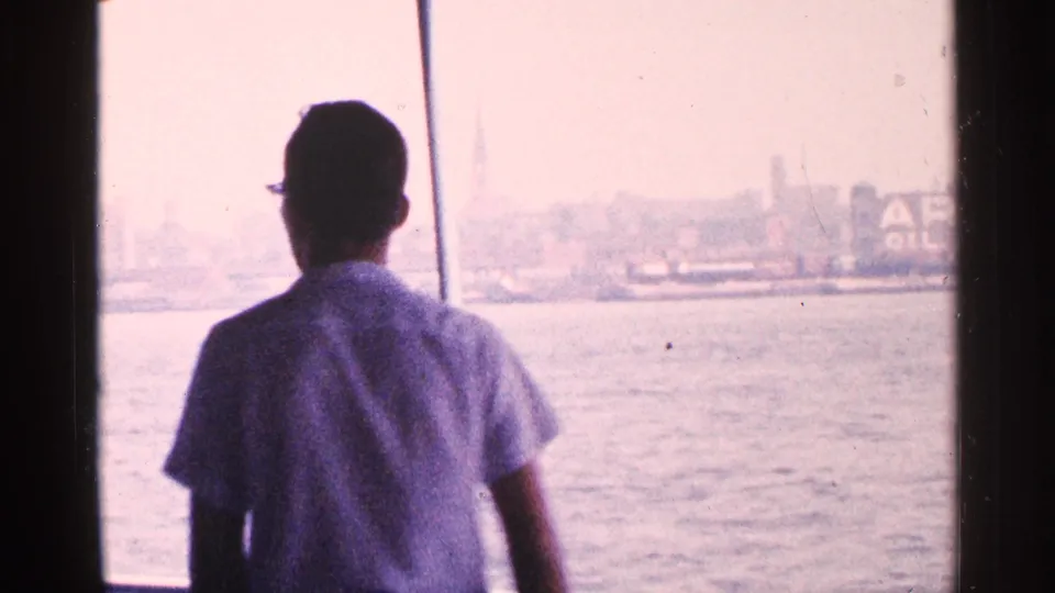 A man in a light shirt stands inside a New York City tour boat, looking out through windows toward the bright waterfront.