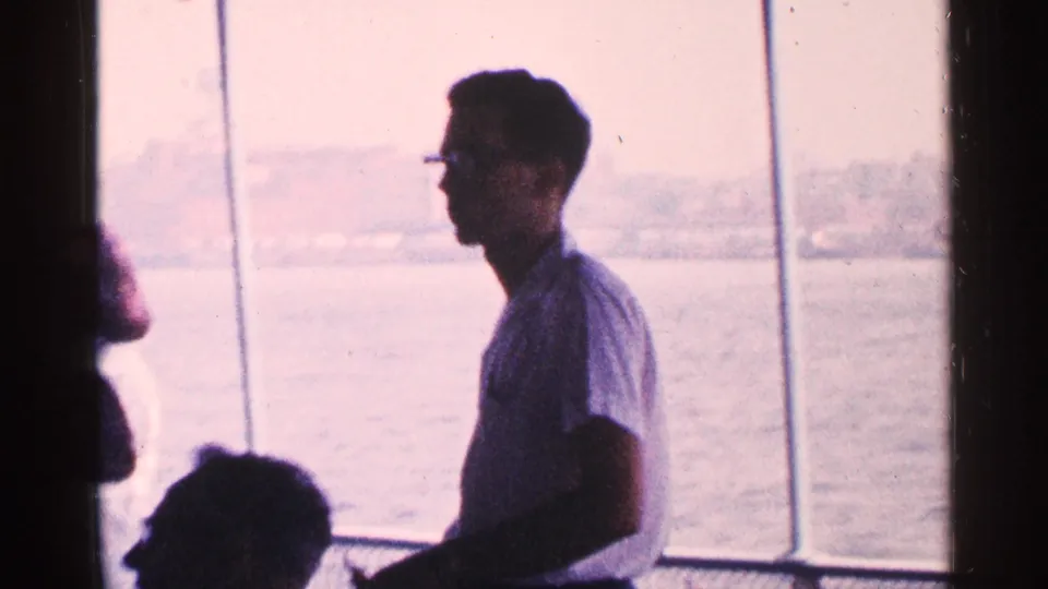 A man wearing glasses is seen in profile on a New York City boat tour, with another passenger and the waterfront beyond the windows.