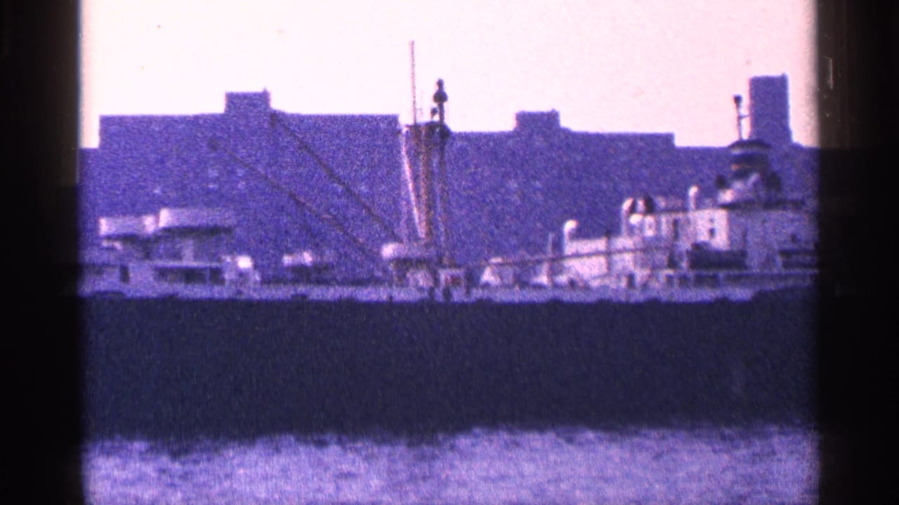 Large ship crossing harbor water in front of dark waterfront buildings.
