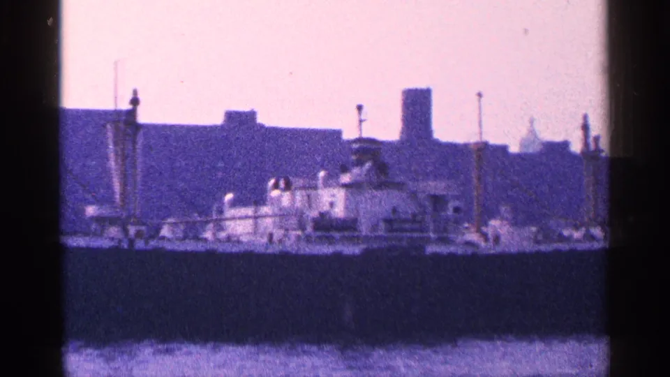 The ship remains in view against New York City waterfront buildings, with harbor water in the foreground and aged color cast.