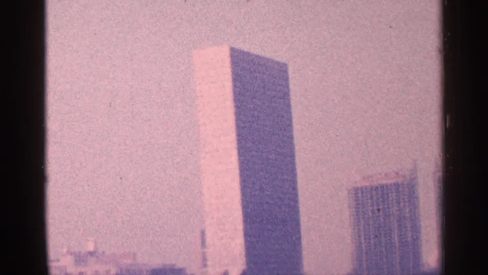 1964 New York City frame showing the United Nations Secretariat Building with a neighboring tower and hazy skyline in 8mm home-movie color.
