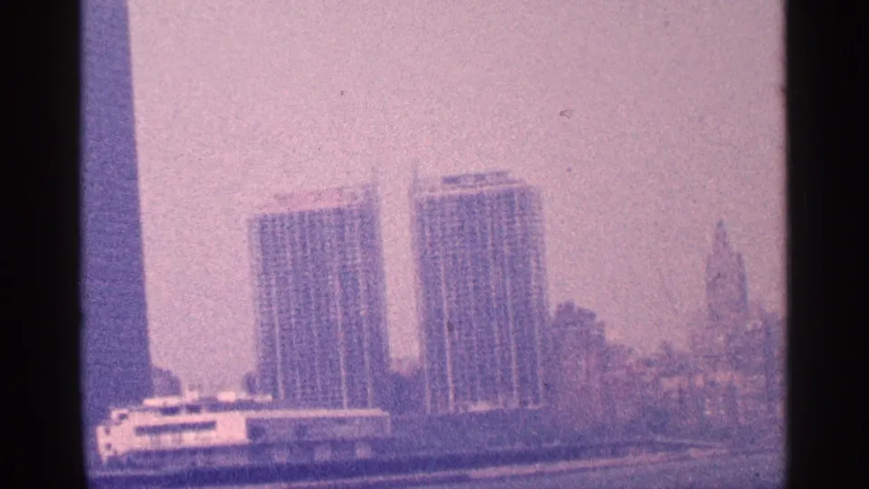 1960s New York City skyline view with waterfront high-rises, a low structure, haze, and visible 8mm frame borders.