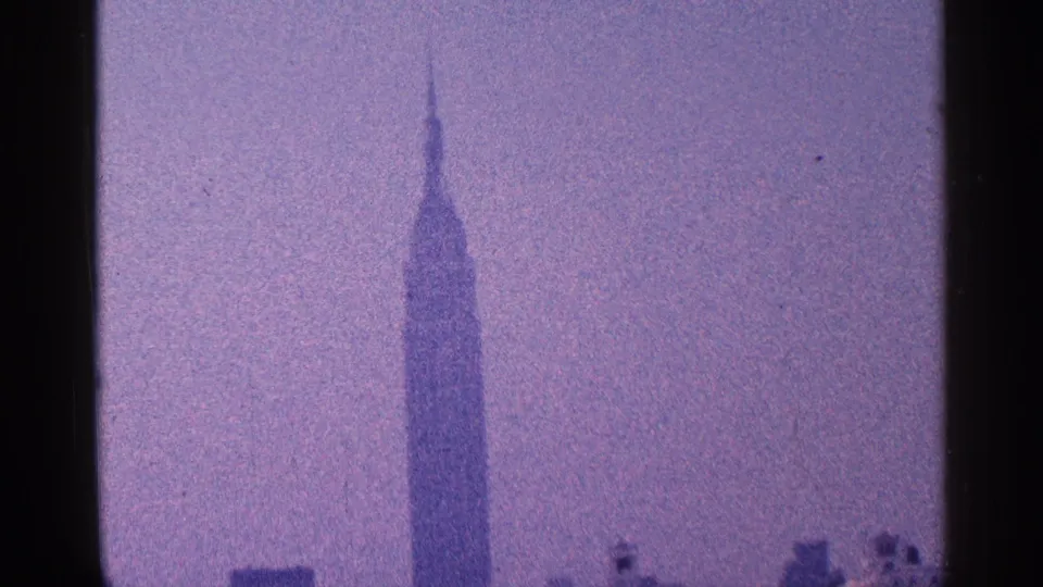 1960s New York City home-movie frame showing the Empire State Building spire against a hazy, blue-purple sky.