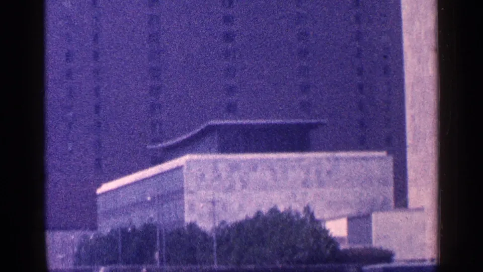 1964 New York City frame showing a low modernist building, trees, and the Empire State Building in the background haze.