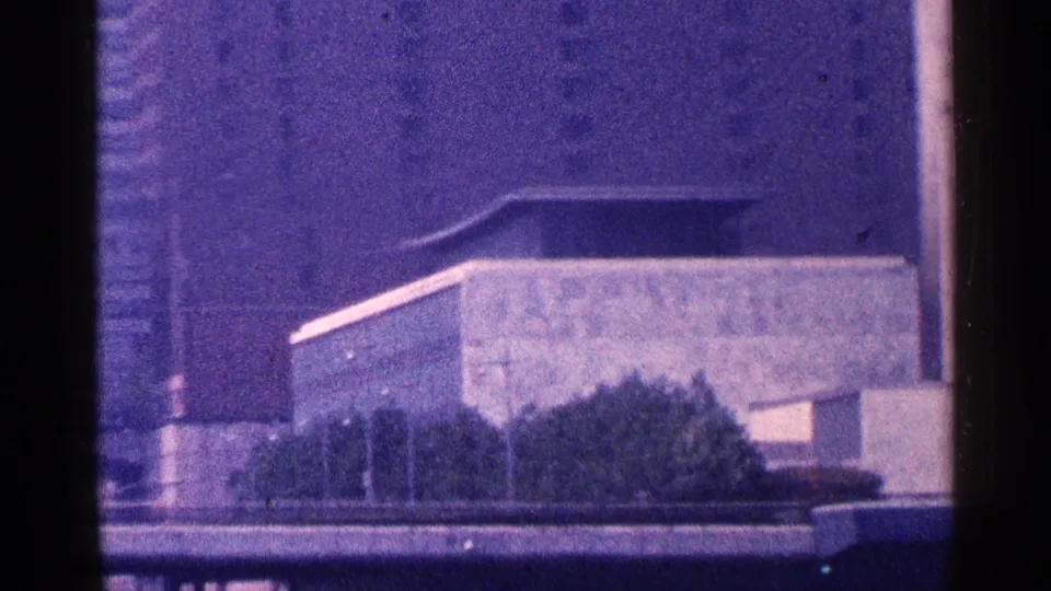 Grainy 8mm view of a modern flat-roofed building and trees with the Empire State Building in the background.