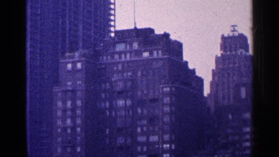 1964 New York City high-rise cluster filmed from an elevated viewpoint, with dark facades, rooftop structures, and heavy 8mm grain.