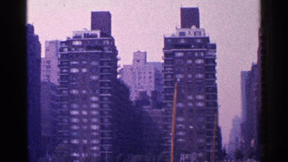 1964 New York City skyline frame with two dark apartment or office towers, rooftop boxes, haze, and a thin pole in the foreground.