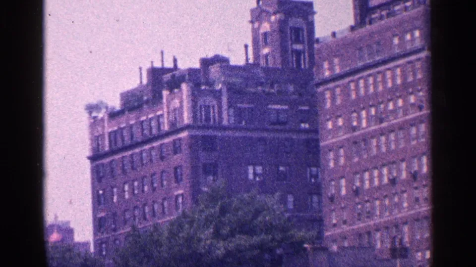 1964 New York City exterior showing older brick residential buildings, roof details, and treetops from a handheld view.