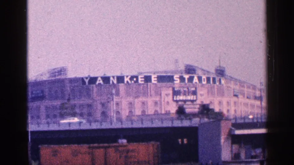1964 home-movie frame of Yankee Stadium in the Bronx, with stadium lettering, facade details, and a vehicle moving below the elevated foreground.