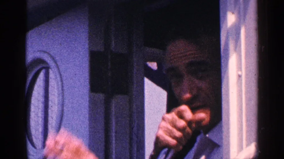 Close-up 1964 home-movie frame of an adult man in a suit standing in a doorway, holding a cigarette and waving toward the camera.