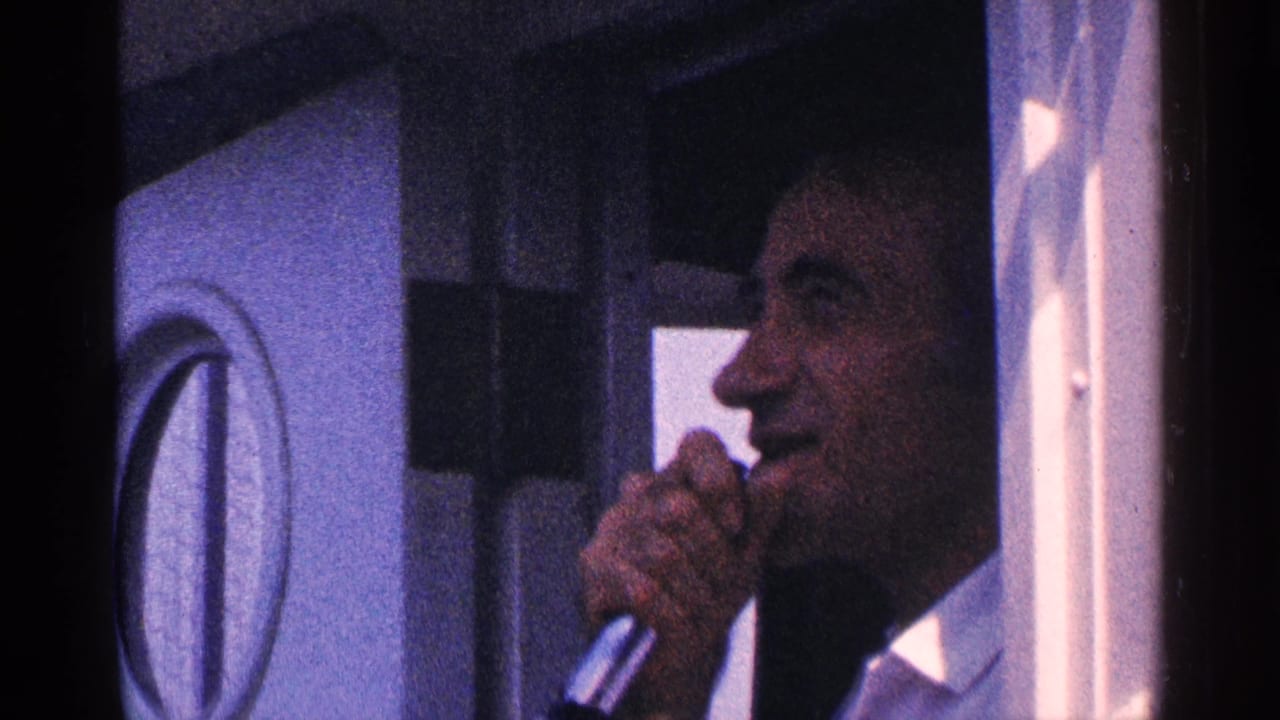 Man holding a microphone inside a vehicle beside a circular window.