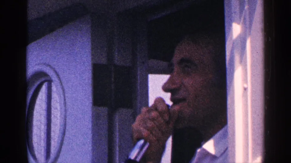 Adult male guide in a light shirt speaks into a handheld microphone inside a vehicle in 1964 New York City footage.