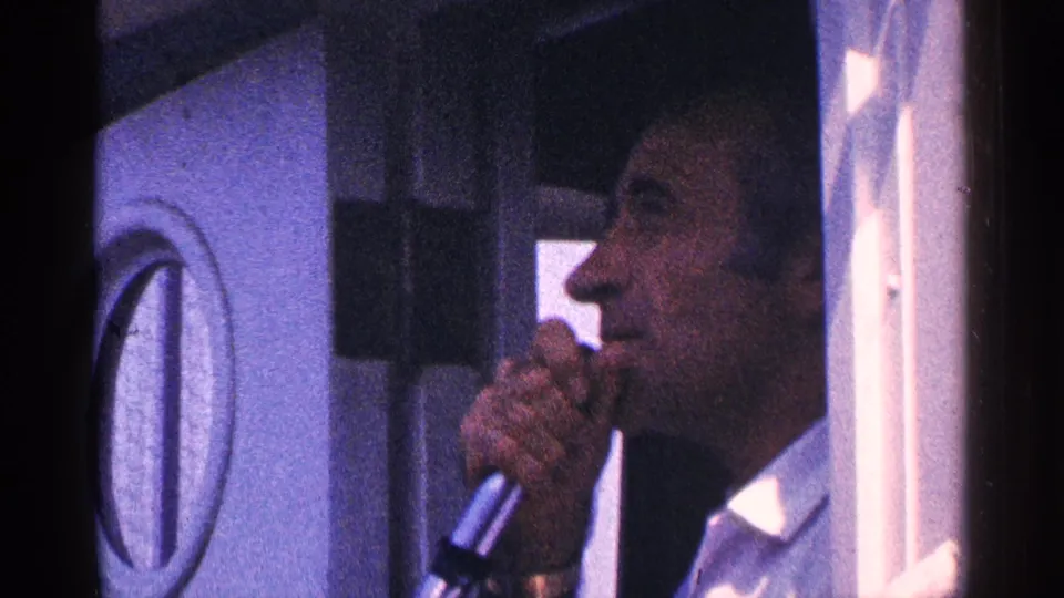 Profile of the adult guide speaking into a microphone beside a circular window in archival 8mm travel footage.