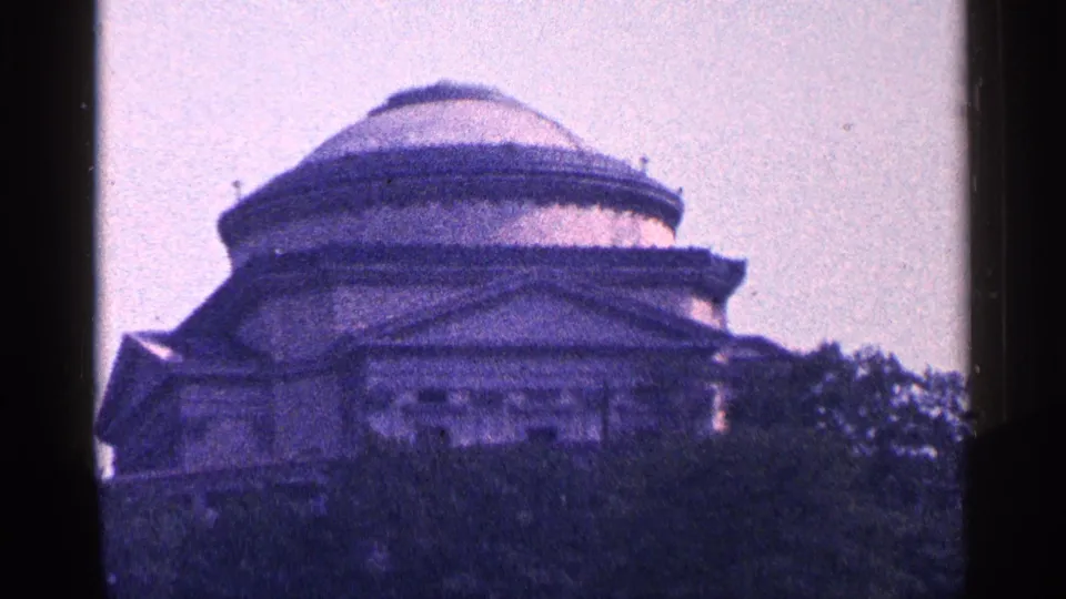 1964 home-movie frame of a distant round-domed building partly hidden by trees, seen in soft purple-tinted 8mm color.