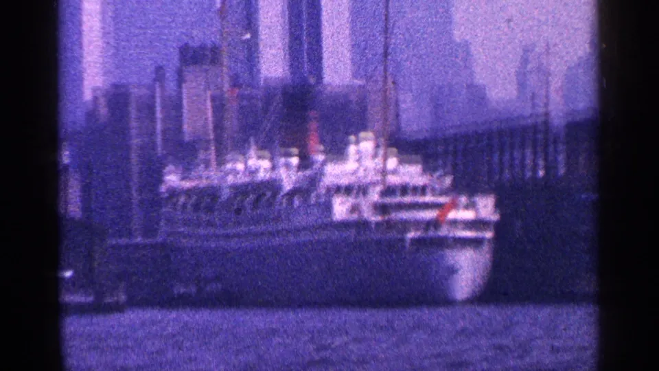 New York City 8mm frame from 1964 showing a passenger ship on the harbor with skyline towers and waterfront structures behind it.