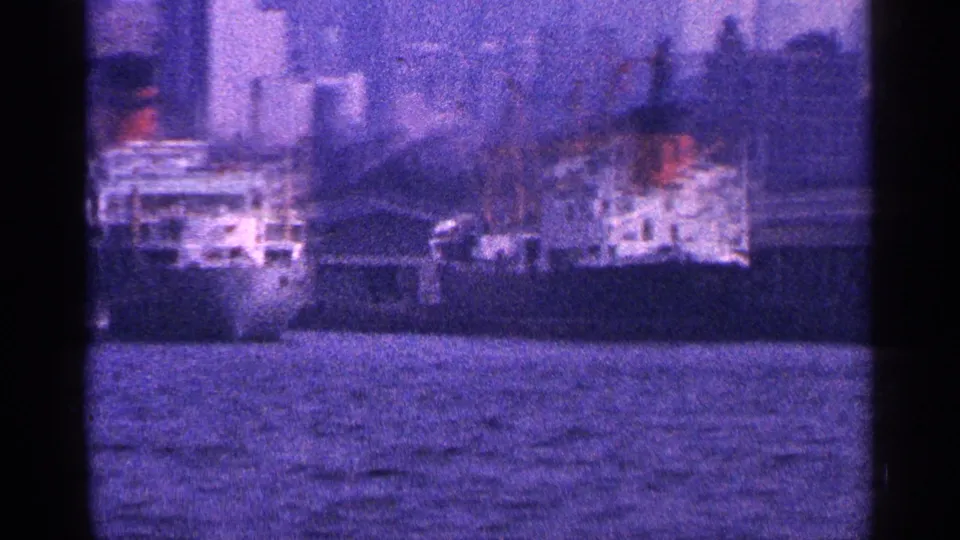 1964 New York City waterfront scene showing moored ships, pier infrastructure, and skyline towers beyond the harbor.