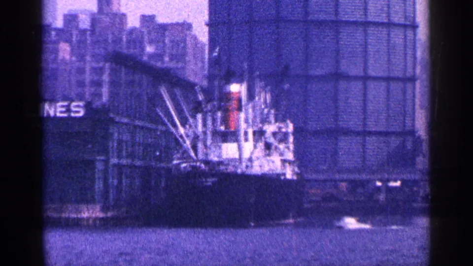 1964 New York City port frame showing a docked vessel, crane equipment, waterfront building, harbor water, and partial signage or lettering.