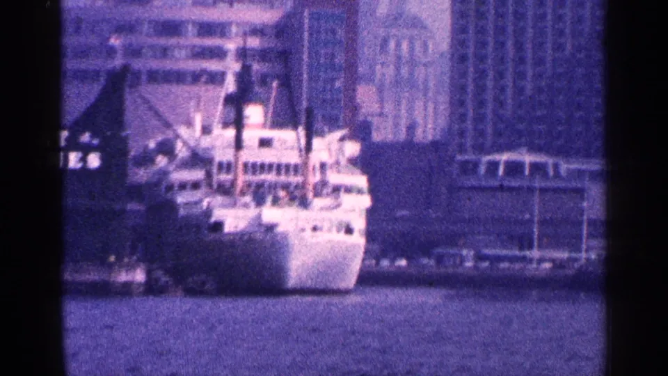 Blue-tinted 8mm frame of a docked white ship in New York City harbor with waterfront buildings and partial commercial signage.