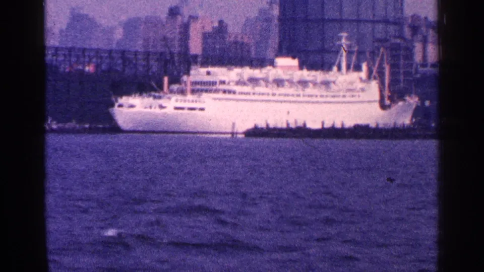 Side view of a white passenger ship along the 1964 New York City harbor, backed by dock structures and hazy skyline buildings.