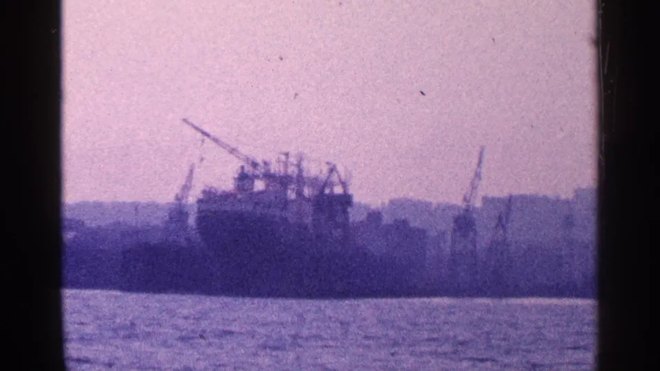 Soft 1964 home-movie frame of New York City waterfront industry with a docked ship, cranes, and harbor water.