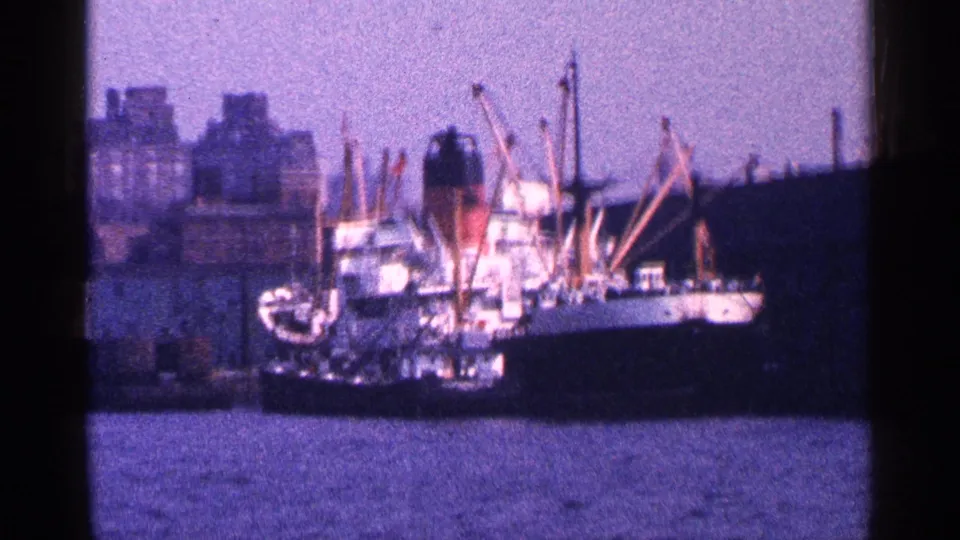 Large cargo ship in New York City harbor, with waterfront buildings, cranes, and water visible in 1964 8mm footage.
