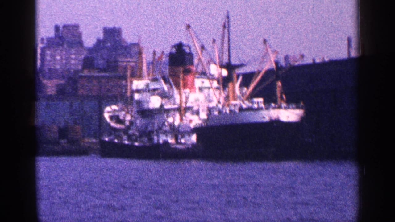 1964 New York harbor view of a large docked cargo ship seen across the water with city buildings and cranes behind it.