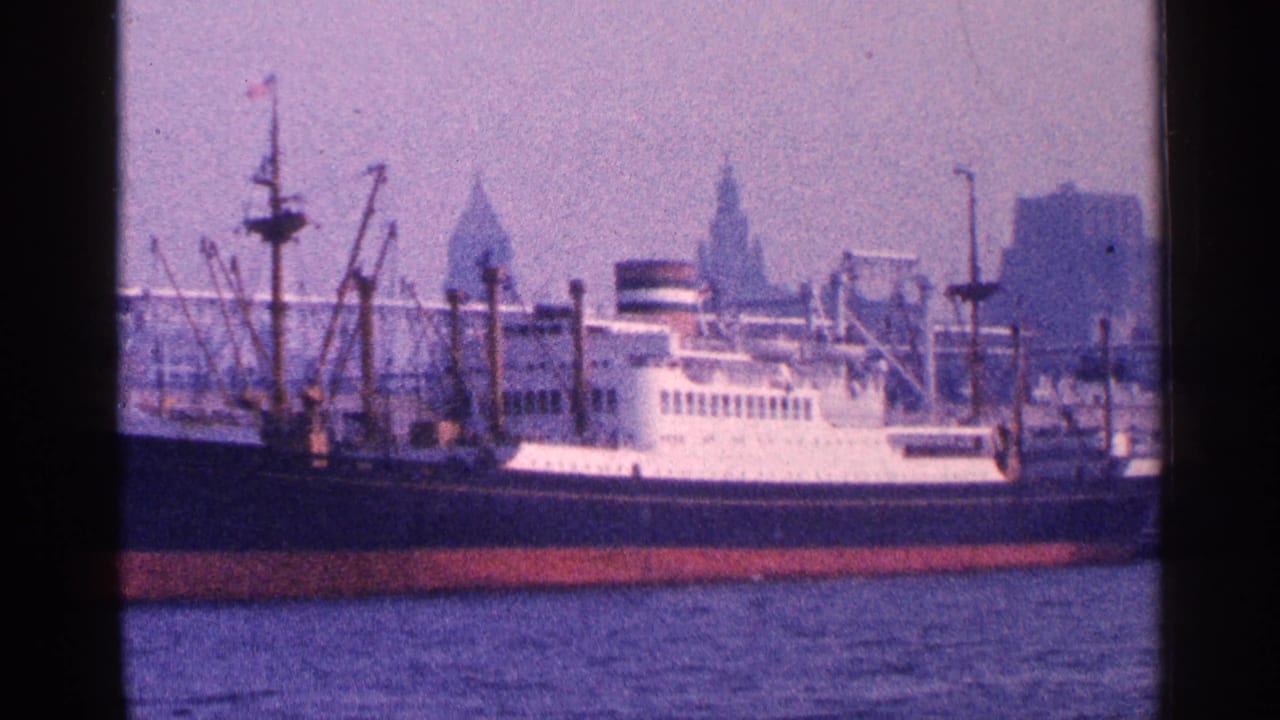 Large docked vessel in New York harbor, with a white superstructure, red lower hull, cranes, and hazy skyline in the background.