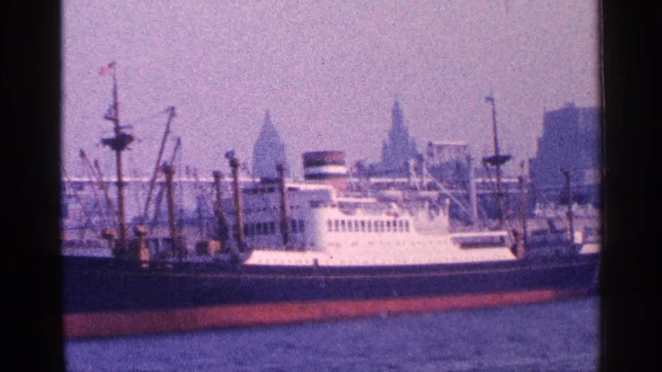 Mid-century New York waterfront view of a docked ship with cranes, water, and distant high-rise silhouettes behind the pier.