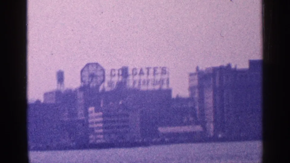 1964 New York City waterfront view with the Colgate Clock and Colgate signage seen across the water on grainy 8mm film.