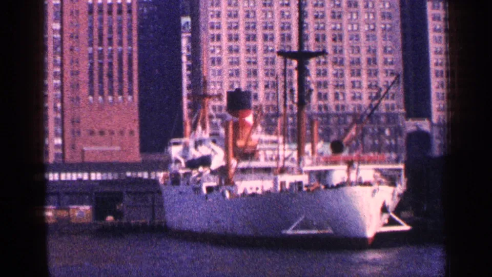 1964 New York City harbor view of a docked white ship with people on deck and tall buildings rising behind the pier.