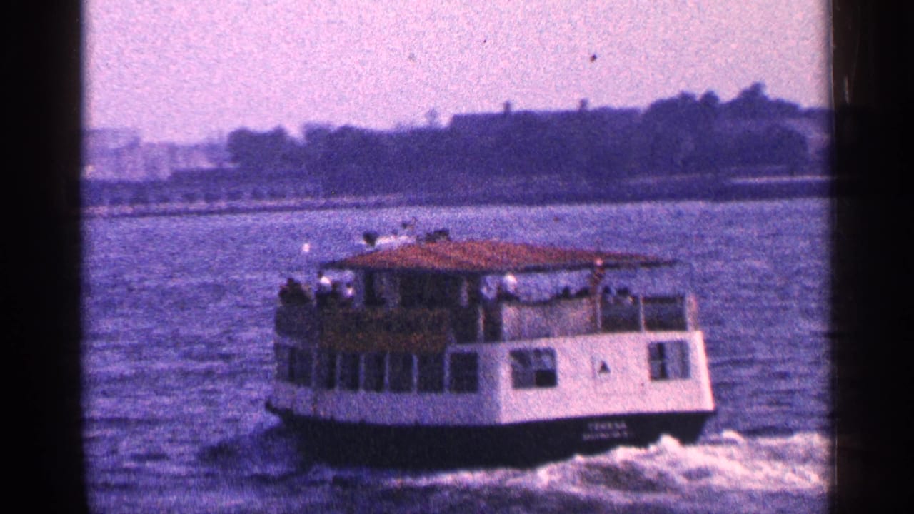 1964 New York City harbor view of a passenger ferry traveling across water, seen in soft 8mm color with distant waterfront silhouettes.
