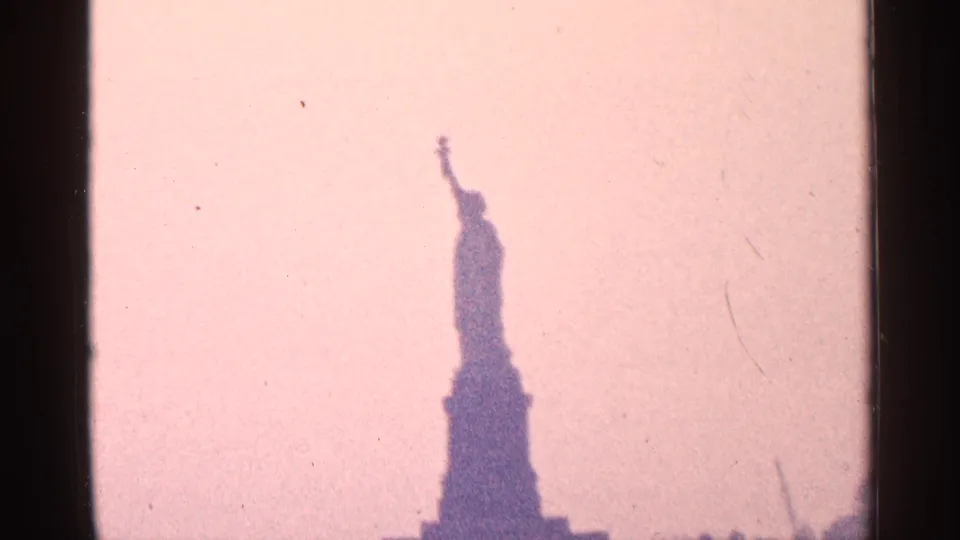 Distant 1964 home-movie view of the Statue of Liberty in silhouette, framed low against a pale pink 8mm sky.