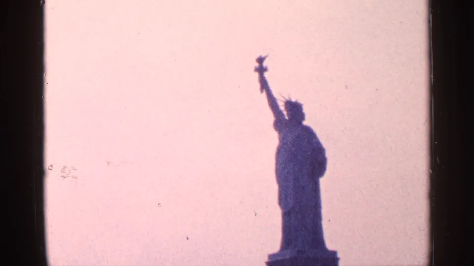 1964 New York City home-movie frame showing the Statue of Liberty silhouetted, with torch and crown visible against pale pink sky.