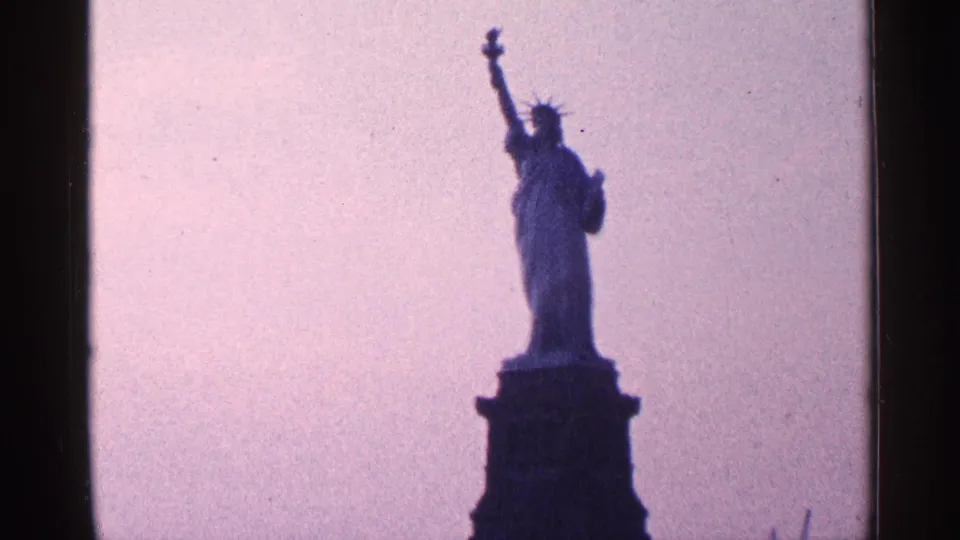 1964 8mm New York City home-movie frame showing the Statue of Liberty in silhouette on its pedestal against a softly colored sky.