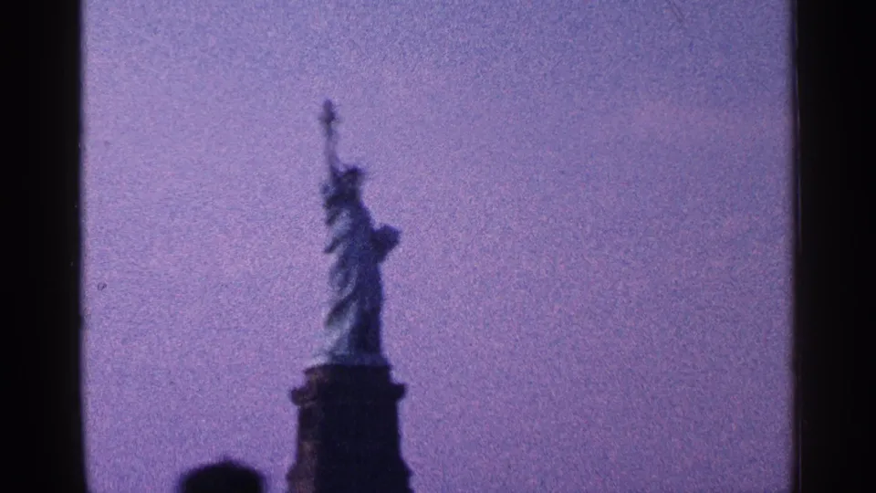 1964 8mm New York City home-movie frame showing a more distant view of the Statue of Liberty and pedestal against a bluish-purple sky.