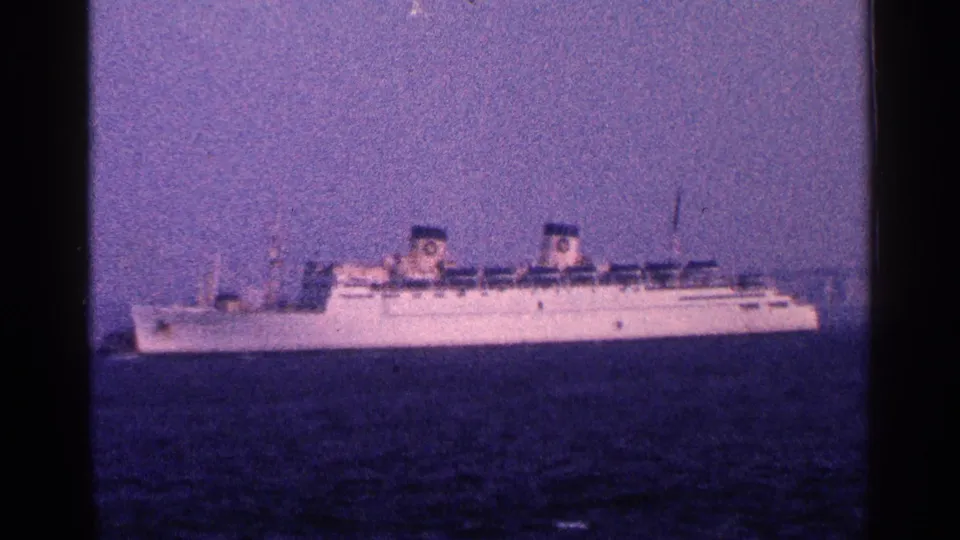 1964 New York City harbor frame showing a large white ocean liner with twin funnels crossing the water in soft, grainy 8mm color.