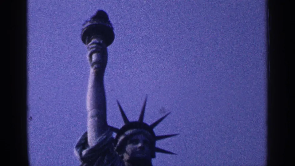 1964 New York City home-movie frame with the Statue of Liberty torch, arm, and crown held against a grainy purple sky.