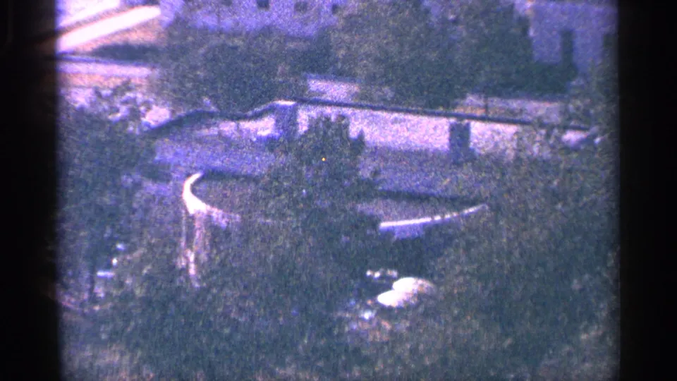 1964 8mm frame with a soft, purple-tinted outdoor view of vegetation, a curved structure, and indistinct waterfront surroundings.
