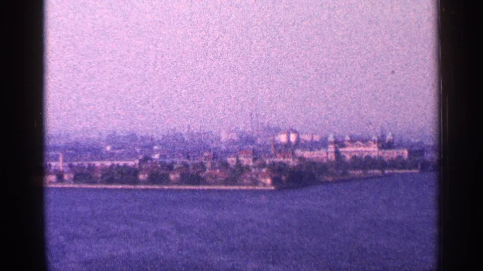 Hazy 1964 New York City skyline view across water, with distant buildings and waterfront trees softened by 8mm grain.