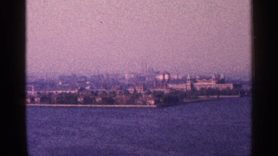 Wide 1964 New York City waterfront view across open water, with a soft skyline and trees under a pale sky.