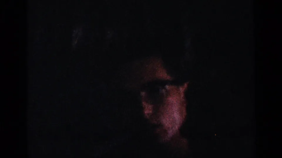 1964 New York home-movie frame showing a man's face partly visible in darkness, with glasses catching limited light.