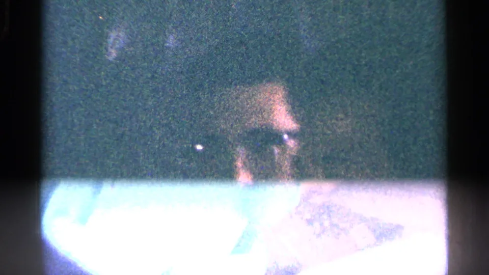 Grainy 8mm frame showing a partially visible adult with glasses, obscured by a bright overexposed horizontal area.